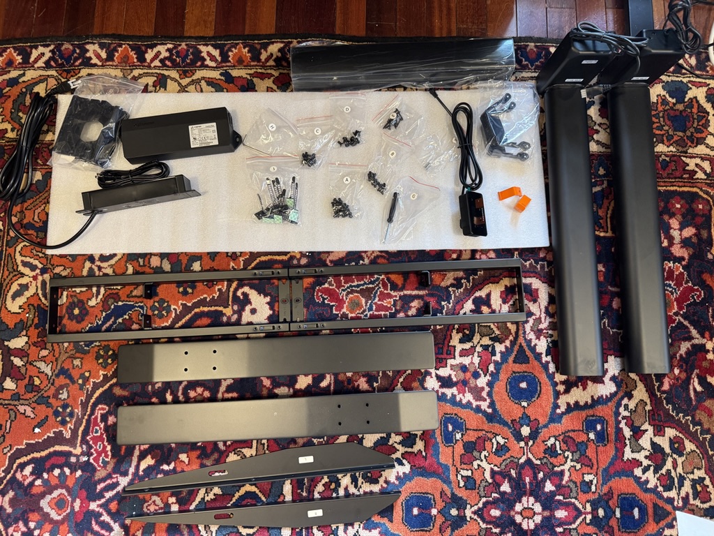 All Autonomous Desk Pro parts laid out on rug: legs, crossbar, frame pieces, labeled hardware bags, motor/control box, power brick, cable management tray, orange headphone hooks