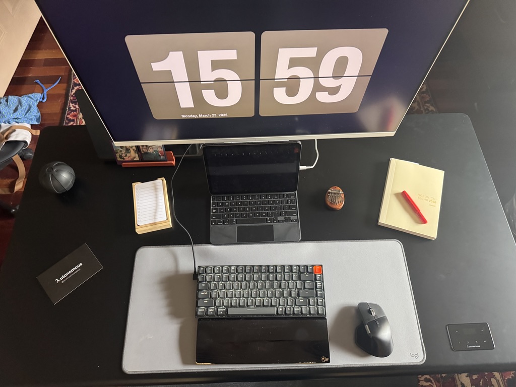 Autonomous Desk Pro full workspace setup from above, showing large monitor with flip clock, iPad, Keychron keyboard, Logitech MX Master mouse, desk mat, and glass controls